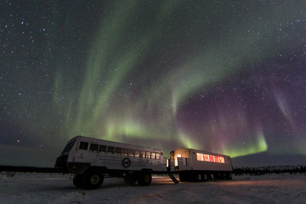 A Tundra Buggy&reg; sits on the icy tundra with an active aurora overhead.