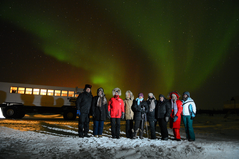 A group of guests stand under the northern lights near a Tundra Buggy.