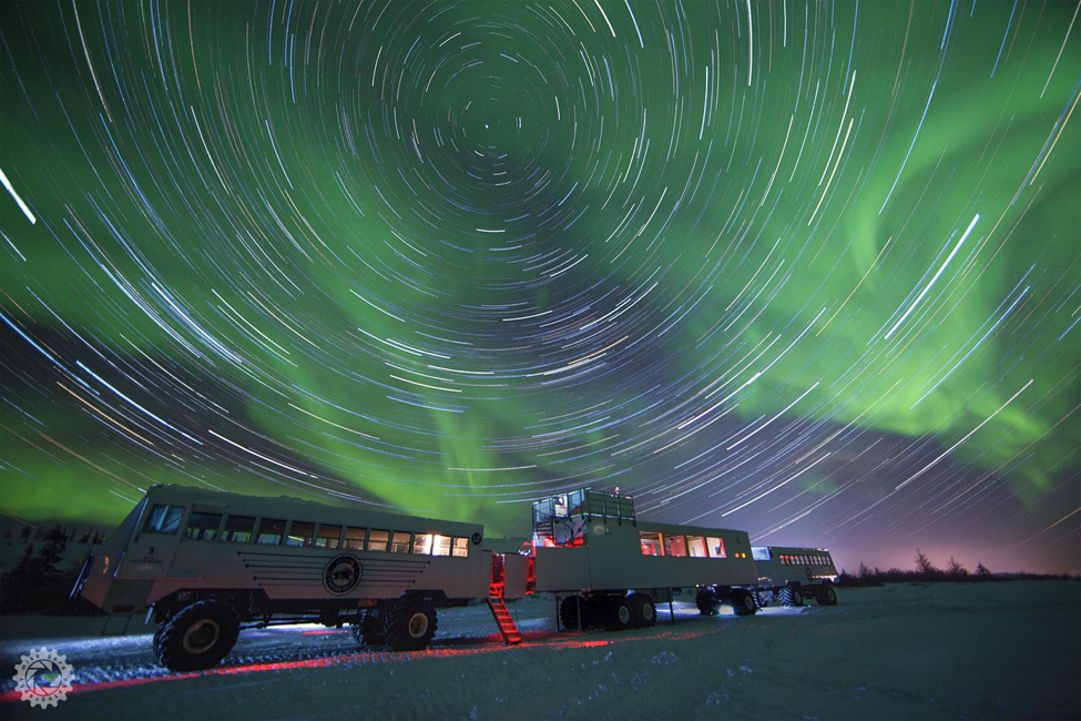 Two Tundra Buggies sit on the tundra under a brilliant northern lights display