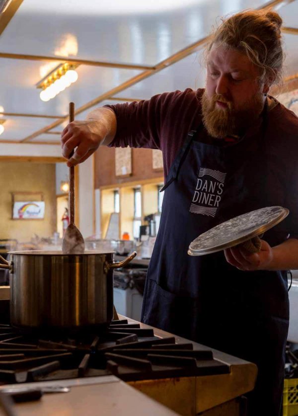 Chef Jared Fossen works in the kitchen, stirring a large pot while wearing a Dan's Diner apron.