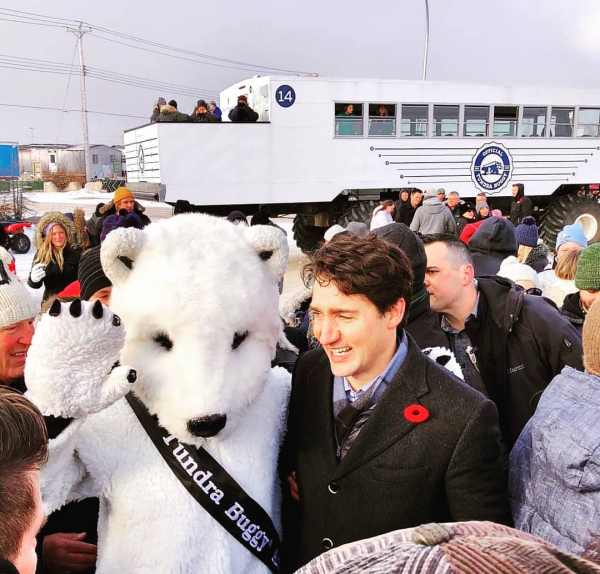 Justin Trudeau stands smiling amongst a crowd with his arm around a person in a bear costume. A Tundra Buggy&reg; sits on the road in the background.